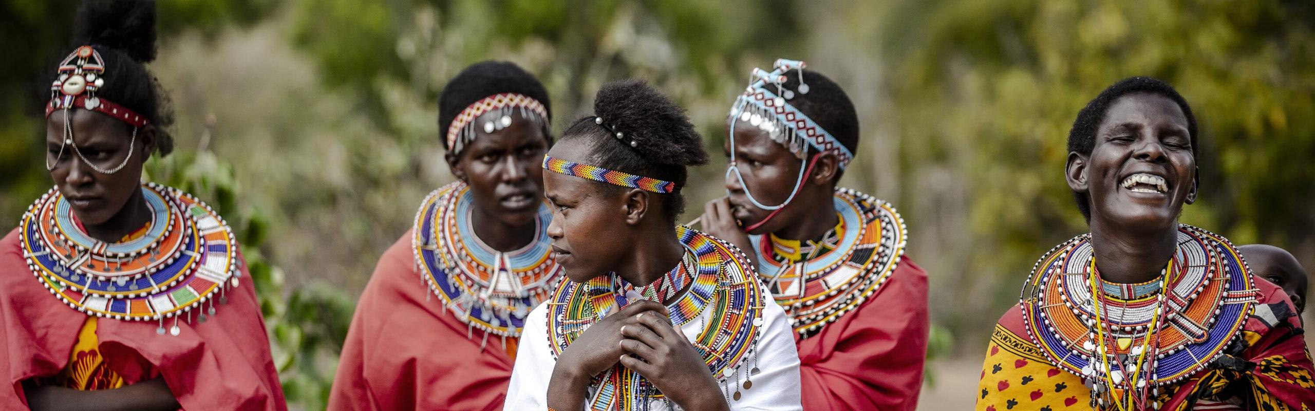 Five members of the Masai community stand together wearing brightly colored traditional clothing and laughing.